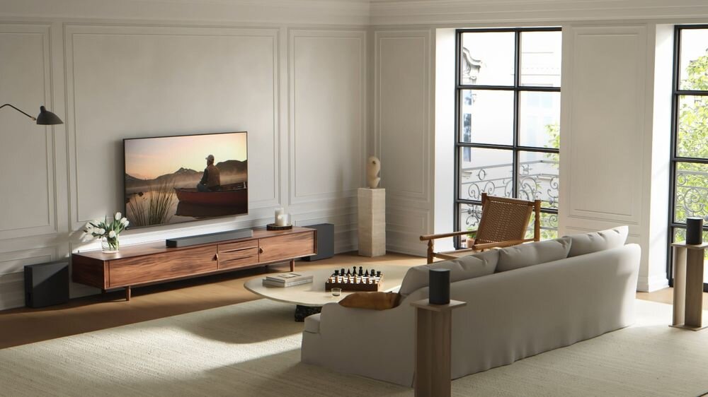 W eleganckim salonie znajduje się duży telewizor na drewnianej szafce, obok którego umieszczono soundbar i głośniki. Przed sofą stoi okrągły stolik z szachami, a przez duże okna wpada naturalne światło.