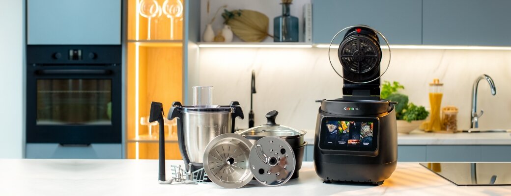 Na blacie kuchennym znajduje się wielofunkcyjny robot kuchenny z otwartą pokrywą, obok którego leżą różne akcesoria, takie jak miska, tarcze i mieszadła. W tle widoczna jest nowoczesna kuchnia z piekarnikiem i zlewem.