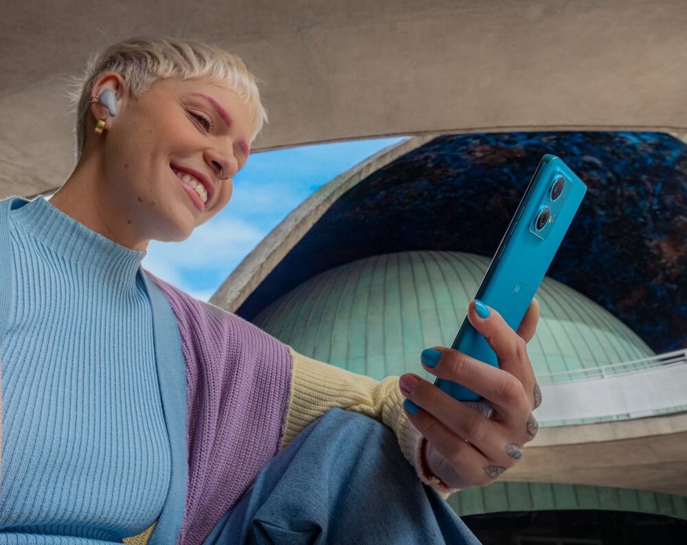 Osoba trzyma niebieski smartfon z dwoma widocznymi aparatami na tylnej obudowie. W tle znajduje się nowoczesna architektura z kopułą.