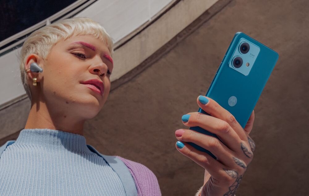 Osoba trzyma w ręku niebieski smartfon z widocznym logo producenta. W tle widać fragment architektury.