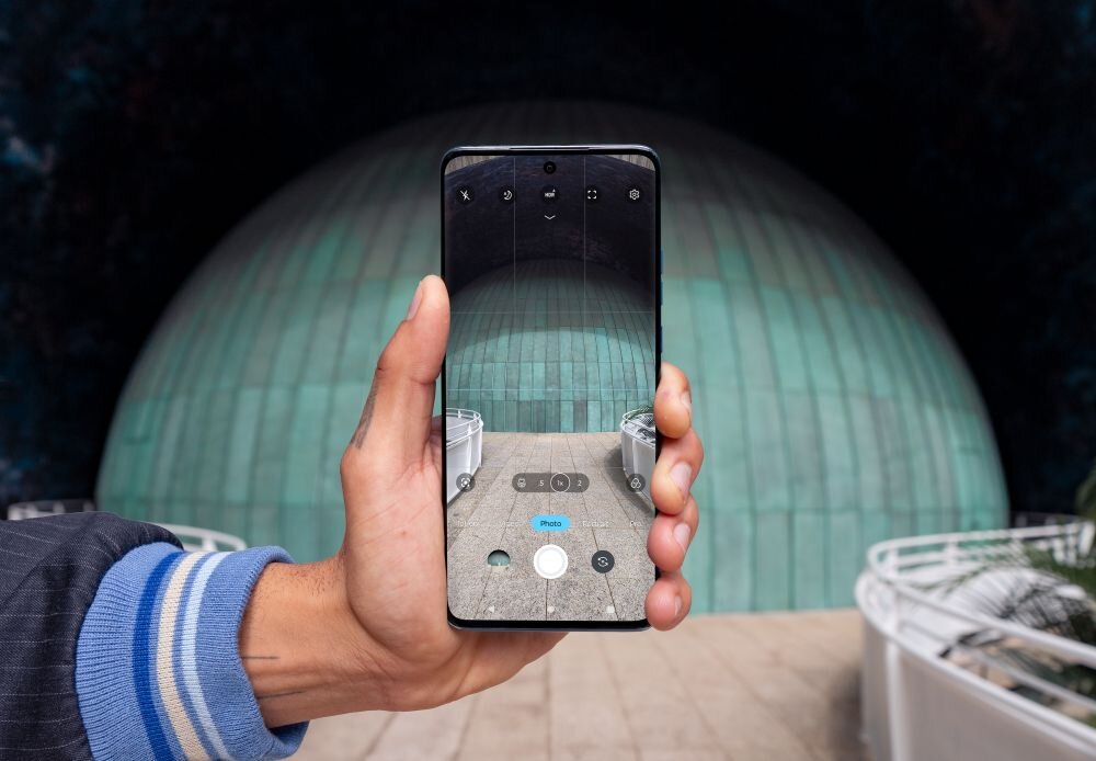 Osoba trzyma smartfon, na ekranie którego widać interfejs aparatu fotograficznego. W tle znajduje się duża, zielona kopuła budynku.