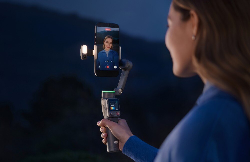Moduł wielofunkcyjny DJI do Osmo Mobile 8P Kobieta trzyma w dłoni szary stabilizator do telefonu skierowany w swoją stronę. Na urządzeniu zamocowany jest smartfon, a obok niego znajduje się mały, świecący moduł, opis