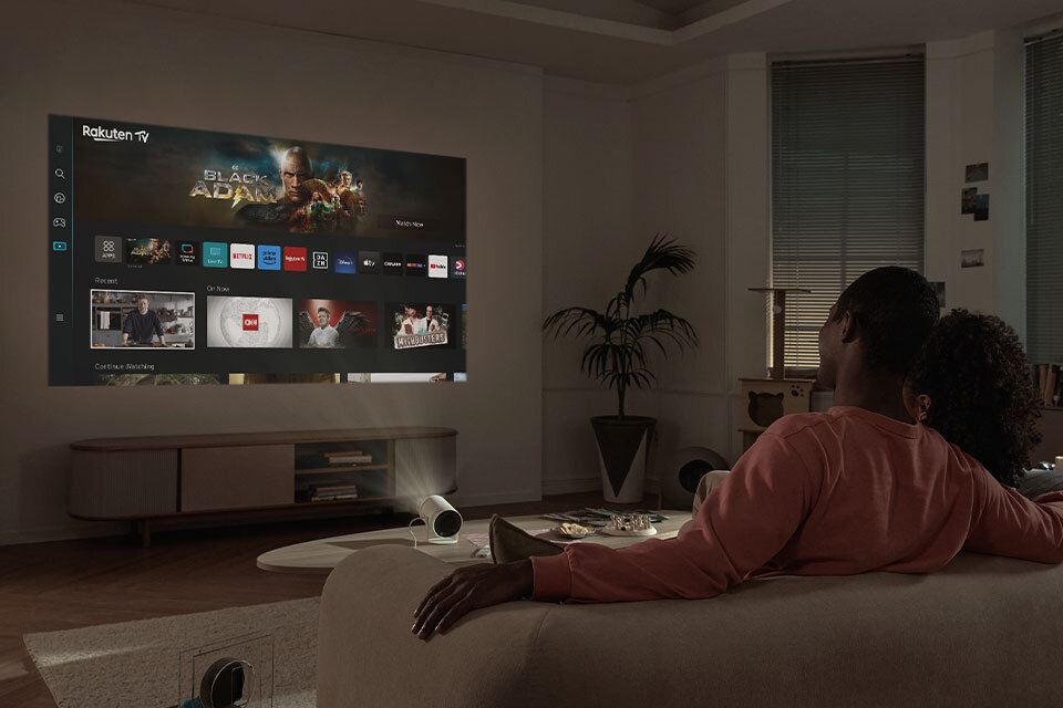 W salonie na ścianie wyświetlany jest obraz z projektora, przedstawiający interfejs Smart TV z aplikacjami takimi jak Rakuten TV i Netflix. Na kanapie siedzą dwie osoby, oglądając projekcję.