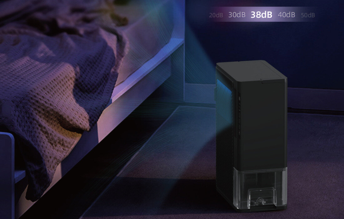 Na obrazie widać czarny osuszacz powietrza stojący na podłodze obok łóżka. W prawym górnym rogu widoczny jest tekst: \'20dB 30dB 38dB 40dB 50dB\'.