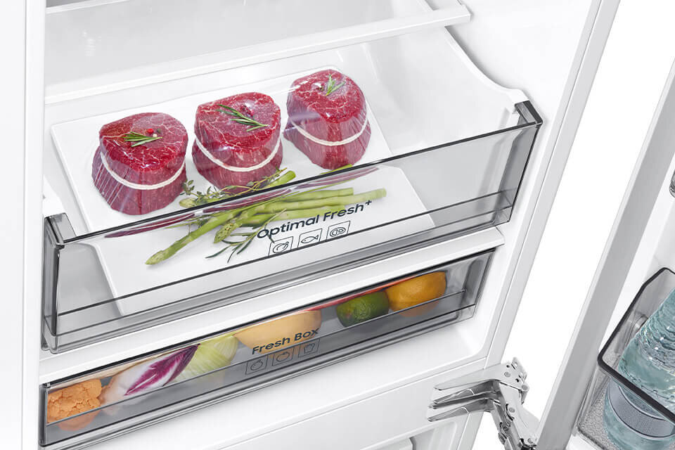 Wnętrze lodówki z szufladą oznaczoną jako \'Optimal Fresh\', w której znajdują się trzy kawałki mięsa i kilka szparagów. Poniżej widoczna jest szuflada \'Fresh Box\' z różnymi warzywami i owocami.
