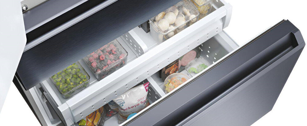 Otwarta szuflada lodówki z widocznymi produktami spożywczymi, takimi jak mrożone warzywa, owoce i grzyby. Wnętrze lodówki jest jasne, a front wykonany z ciemnego szkła.