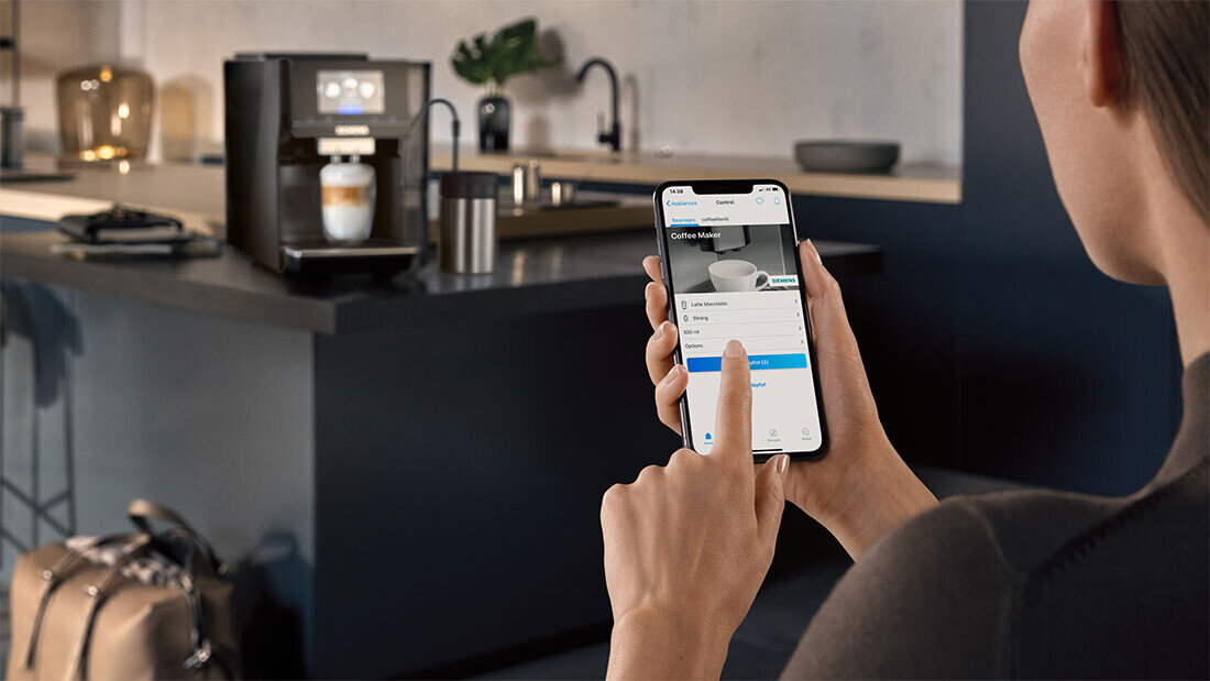 Osoba trzyma smartfon z aplikacją do sterowania ekspresem do kawy, na ekranie widoczny jest napis \'Coffee Maker\'. W tle znajduje się ekspres do kawy z filiżanką pod dyszą.