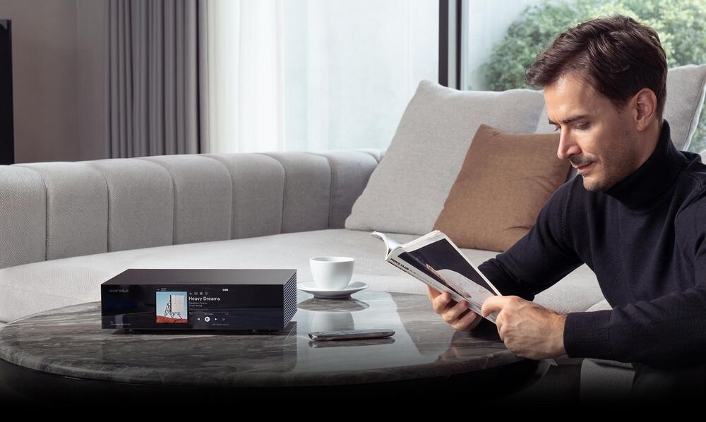 Mężczyzna siedzi na kanapie i czyta książkę, obok na stoliku znajduje się czarny sprzęt audio z wyświetlaczem pokazującym okładkę albumu i tekst \'Heavy Dream\'.