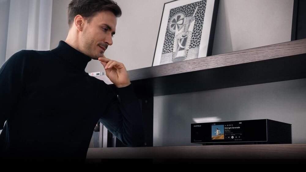 Mężczyzna stoi obok czarnego urządzenia audio z wyświetlaczem dotykowym, na którym widoczny jest napis \'media expert\'. Na półce znajduje się również oprawiony obrazek.