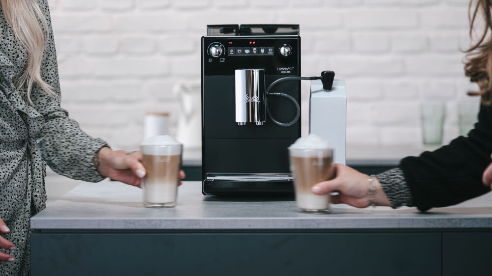 Ekspres do kawy z widocznym logo Melitta stoi na blacie, obok znajduje się pojemnik na mleko. Dwie osoby trzymają szklanki z kawą z mleczną pianką.
