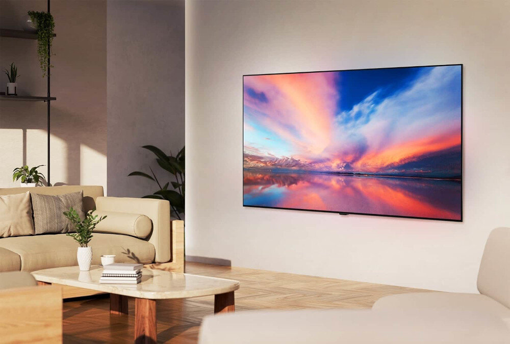 Telewizor zamontowany na ścianie w nowoczesnym salonie, wyświetlający kolorowy krajobraz z zachodem słońca. W pomieszczeniu znajdują się jasne meble i rośliny doniczkowe.