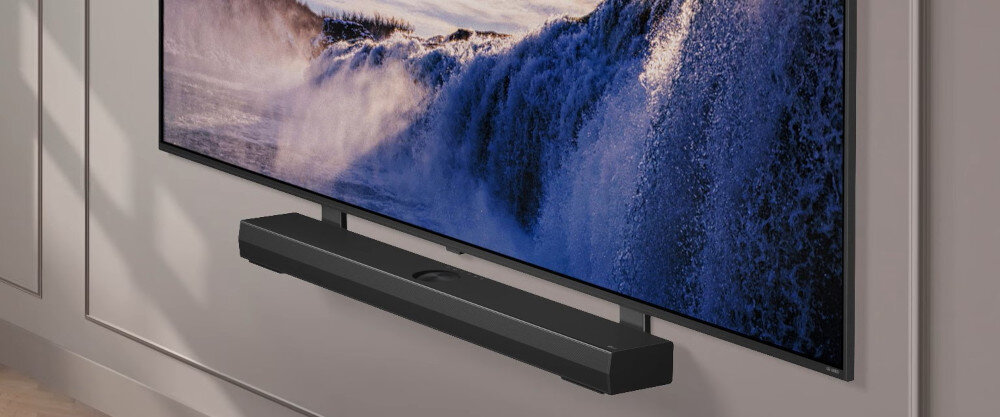 Na obrazie widoczny jest duży telewizor zamontowany na ścianie, wyświetlający obraz fal morskich. Pod telewizorem znajduje się czarny soundbar.