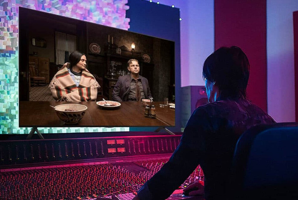 Osoba siedzi przed dużym telewizorem, na którym wyświetlana jest scena z dwiema osobami przy stole. W tle widoczne są elementy studia nagraniowego z kolorowym oświetleniem.