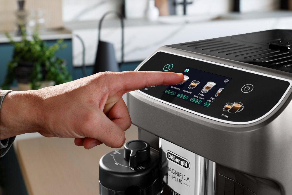 Osoba dotyka panelu dotykowego ekspresu do kawy, na którym widoczne są różne opcje napojów, takie jak Cappuccino, Espresso, Latte. Na obudowie ekspresu znajduje się logo \'DeLonghi\' oraz napis \'Magnifica Plus\'.