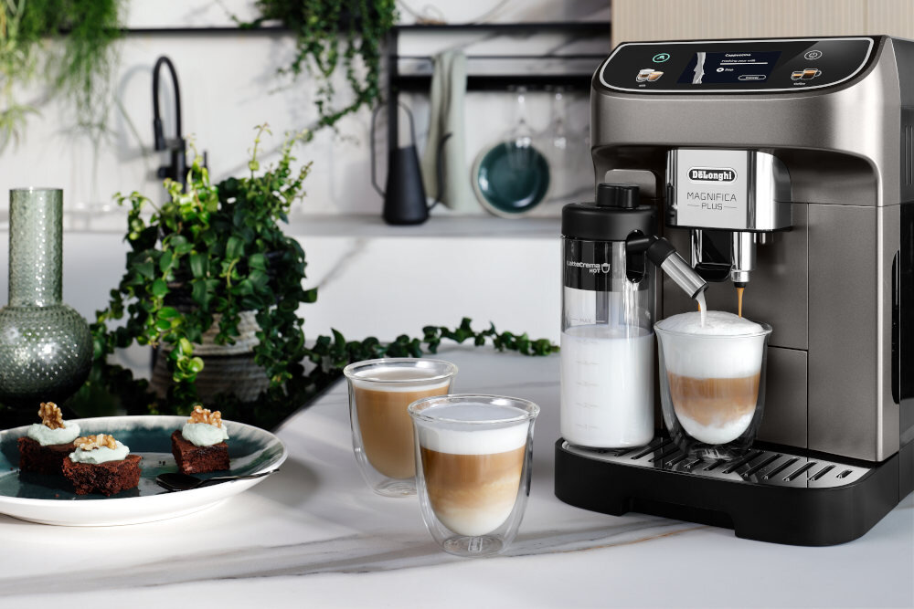 Na blacie kuchennym stoi ekspres do kawy z logo \'DeLonghi Magnifica Plus\', obok którego znajdują się trzy szklanki z kawą i talerz z ciastkami. W tle widoczne są rośliny i elementy dekoracyjne.