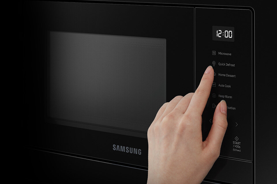 Czarna kuchenka mikrofalowa z wyświetlaczem pokazującym godzinę 12:00 i panelem dotykowym z opcjami: Microwave, Quick Defrost, Home Dessert, Auto Cook, Keep Warm, Start. Logo Samsung widoczne na froncie urządzenia.