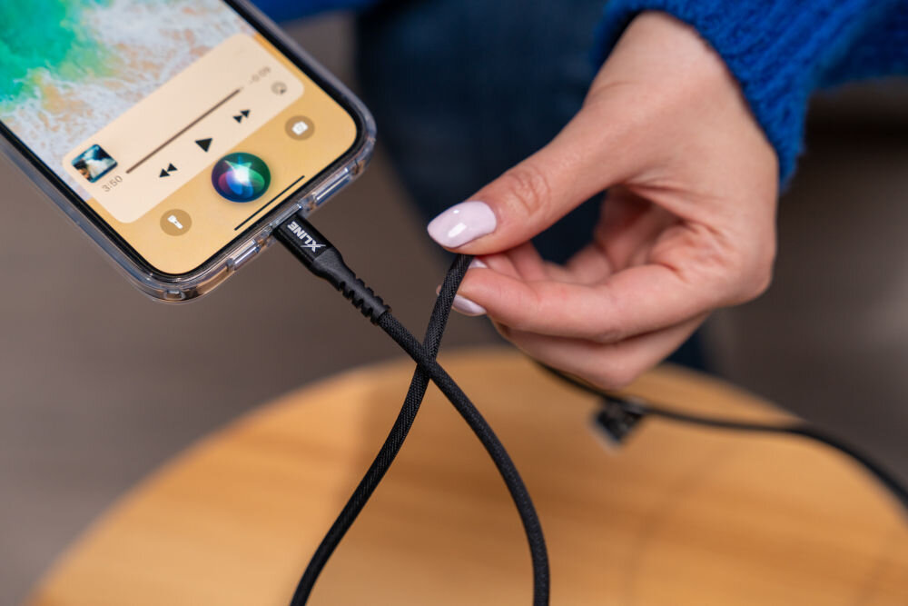 Osoba trzyma kabel USB-C - Lightning podłączony do smartfona, na którym widoczny jest ekran z odtwarzaczem muzyki. Na kablu znajduje się logo \'XLINE\'.