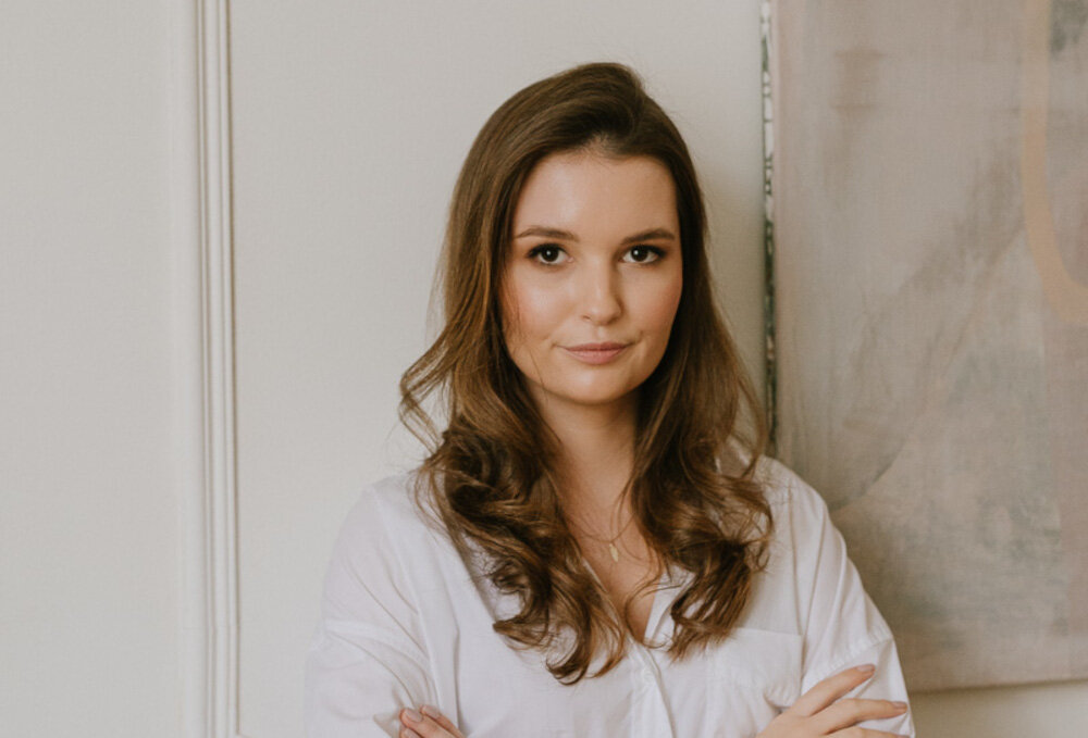 Osoba o długich, falowanych włosach stoi na tle jasnej ściany i obrazu. Ubrana jest w białą koszulę.