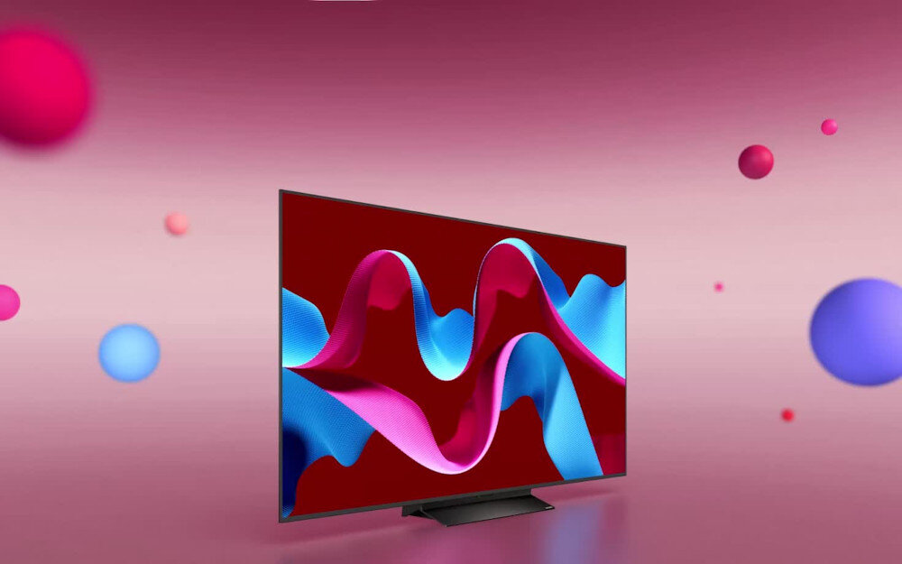 Telewizor z cienką ramką wyświetla kolorową grafikę z falistymi kształtami na ekranie. W tle widoczne są różnokolorowe kule na gradientowym różowym tle.
