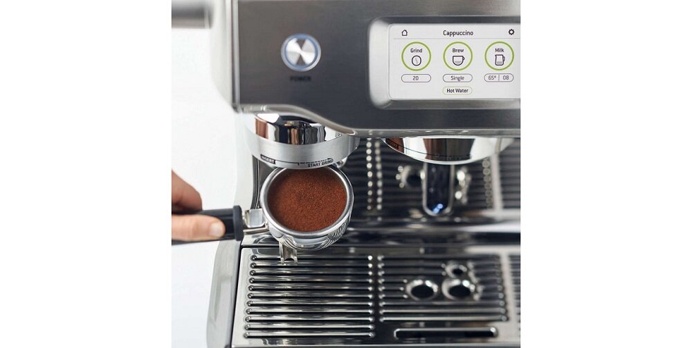 Ekspres do kawy z wyświetlaczem pokazującym opcje \'Cappuccino\', \'Grind\', \'Brew\', \'Milk\' oraz \'Hot Water\'. Widoczny jest portafilter z mieloną kawą umieszczony w ekspresie.