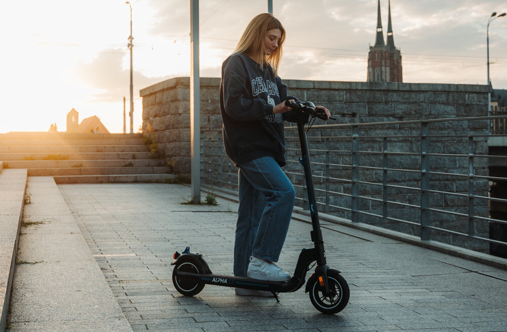 Osoba stoi na czarnej hulajnodze elektrycznej na tle miejskiego krajobrazu o zachodzie słońca. Na hulajnodze widoczny jest napis \'ALPHA\'.