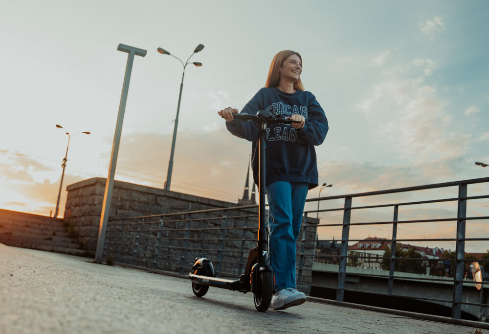 Osoba stoi na chodniku trzymając czarną hulajnogę elektryczną. W tle widać latarnie i fragmenty budynków.