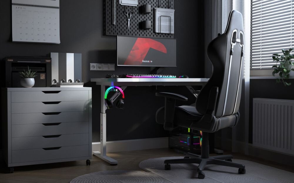 Na obrazie znajduje się biurko gamingowe z białym blatem, na którym stoi monitor i klawiatura z podświetleniem RGB. Obok biurka znajduje się czarne krzesło gamingowe, a w tle widać szafkę z szufladami i kalendarz na ścianie.