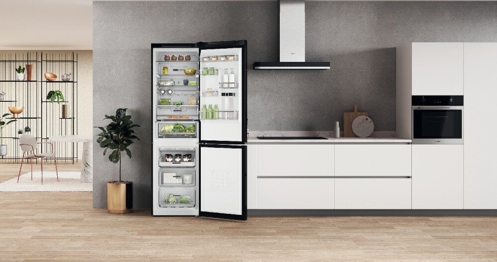 Kuchnia z otwartą lodówką w kolorze czarnym i inox, wypełnioną różnymi produktami spożywczymi. Obok znajduje się biały blat z zlewem, okapem i piekarnikiem w zabudowie.