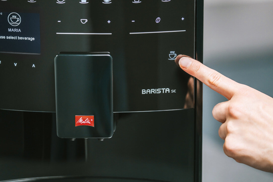 Zbliżenie na panel sterowania ekspresu do kawy z logo Melitta i napisem BARISTA SE. Osoba naciska przycisk z ikoną filiżanki i napisem \'my\'.