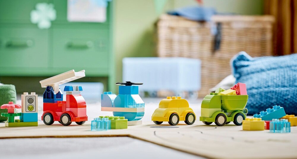 Na obrazku znajdują się kolorowe klocki LEGO DUPLO ułożone w kształty pojazdów, takich jak ciężarówka, helikopter i samochody. W tle widoczne są rozmyte elementy wnętrza pokoju dziecięcego.