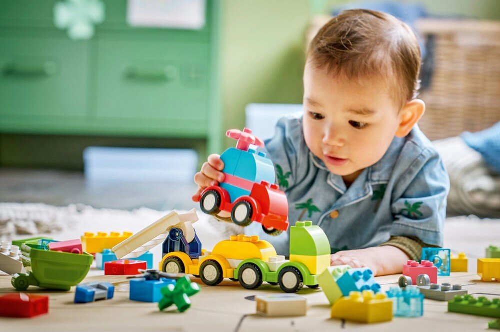 Dziecko bawi się kolorowymi klockami, tworząc pojazdy z zestawu LEGO DUPLO. W tle widoczne są inne elementy zestawu rozłożone na podłodze.