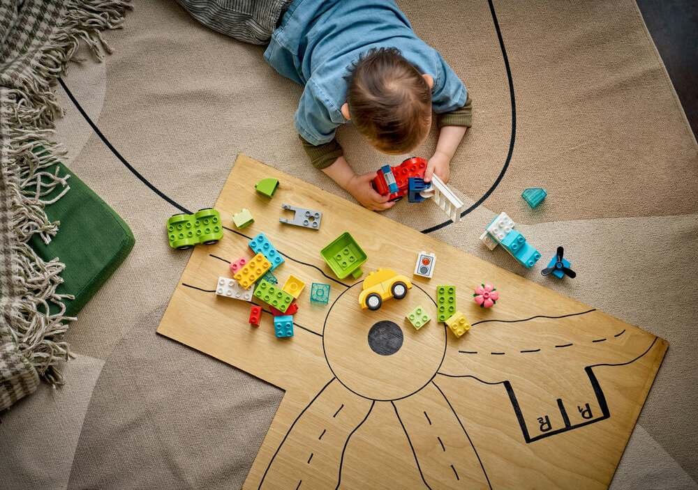 Dziecko bawi się klockami LEGO na drewnianej planszy z narysowanymi drogami. Na planszy znajdują się różnokolorowe klocki i mały żółty samochodzik.