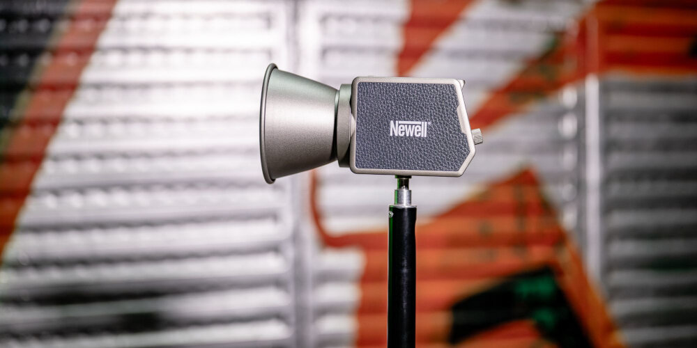 Lampa LED Newell z zamontowaną metalową czaszą, ustawiona na czarnym uchwycie na tle rozmytych regałów magazynowych kv lifestyle początek opisu sekcja nagłówkowa