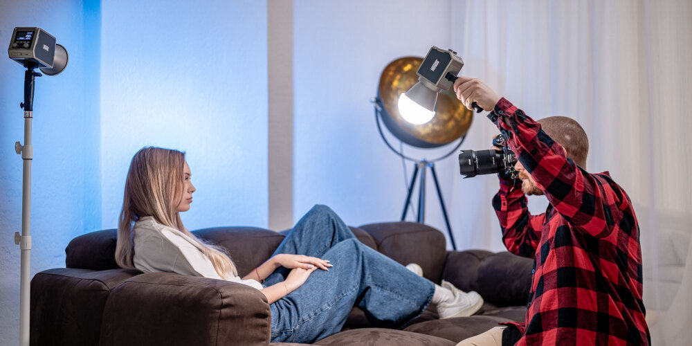 Fotograf wykonuje zdjęcie kobiety siedzącej na kanapie, trzymając lampę LED Newell z czaszą, a druga lampa stoi na statywie