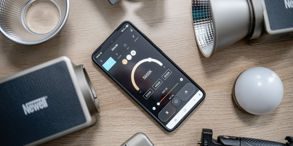 Smartfon z otwartą aplikacją do sterowania lampą LED Newell leży obok lampy, czaszy, silikonowego dyfuzora i uchwytu na drewnianym blacie.
