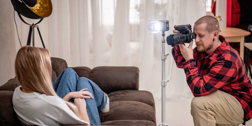 LAMPA LED NEWELL ZORA MINI 40 RGB Fotograf wykonuje zdjęcie osoby siedzącej na kanapie, obok aparatu stoi na statywie świecąca lampa LED z otwartymi wrotami