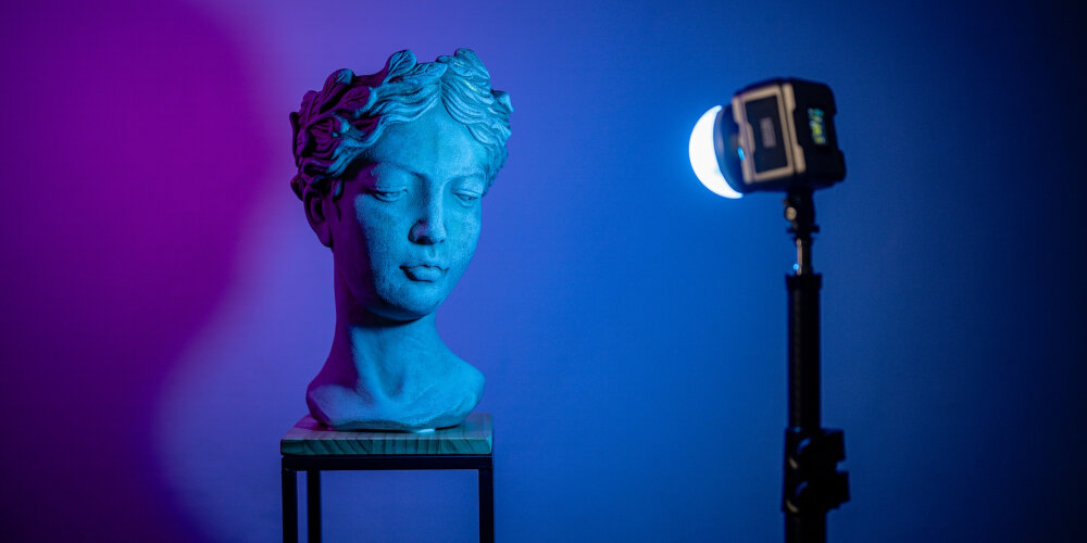 LAMPA LED NEWELL ZORA MINI 40 RGB Popiersie ustawione na stoliku oświetlone niebieskim i fioletowym światłem, obok na statywie świeci lampa LED z dyfuzorem