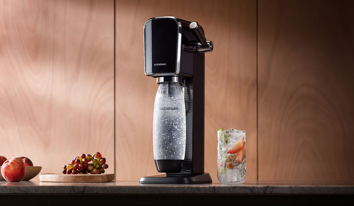 Czarny saturator z logo \'SodaStream\' stoi na blacie obok szklanki z napojem i owocami. W tle widoczne są winogrona i brzoskwinie na drewnianym tle.