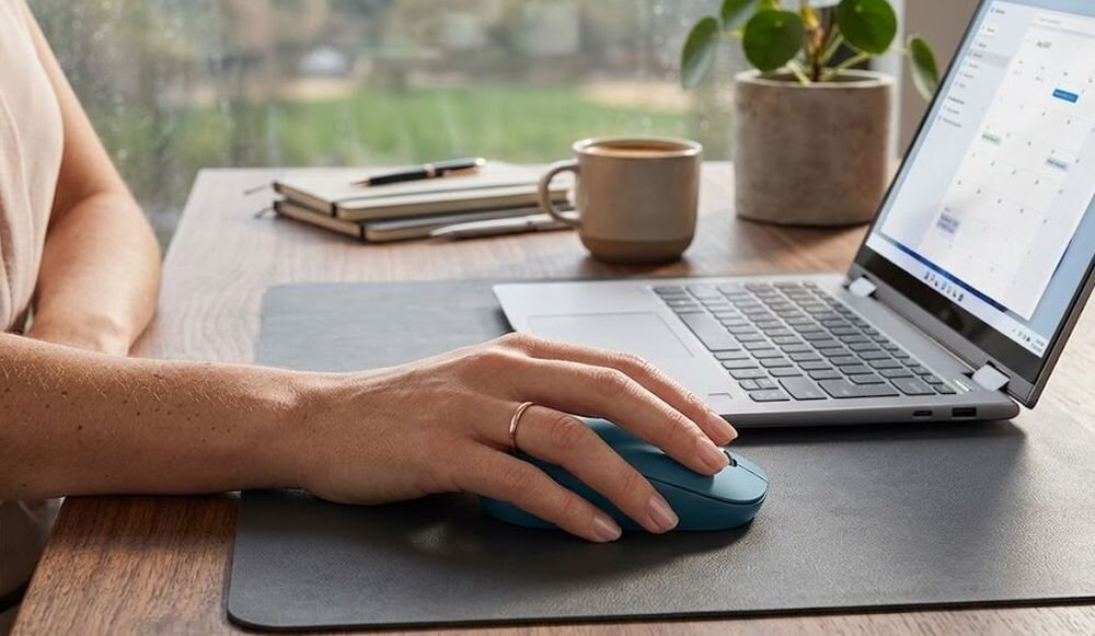 Osoba używa niebieskiej myszy komputerowej przy laptopie na biurku. W tle widać roślinę doniczkową i notatnik.