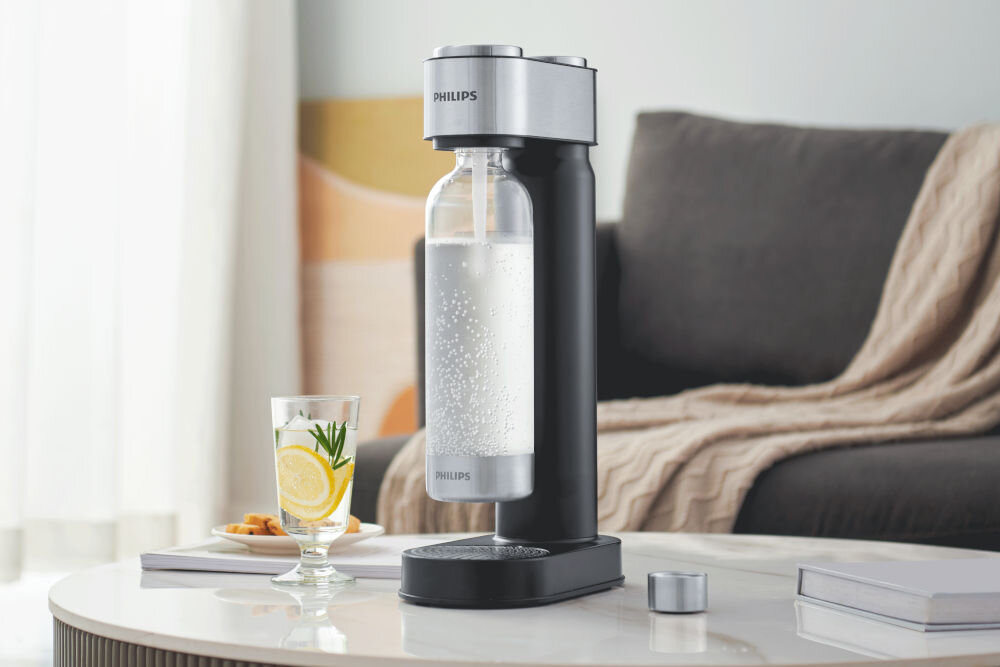 Na stoliku znajduje się czarny saturator marki PHILIPS z przezroczystą butelką wypełnioną wodą gazowaną. Obok stoi szklanka z napojem i plasterkami cytryny oraz gałązką rozmarynu.
