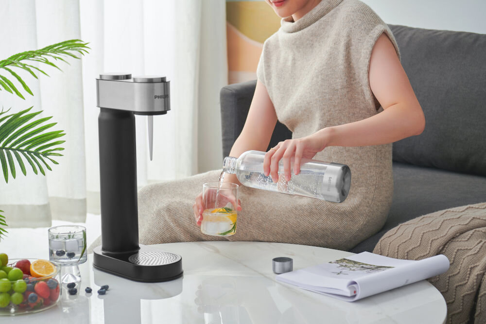 Osoba siedzi na kanapie i nalewa wodę z butelki do szklanki na stoliku, obok stoi czarny saturator z logo PHILIPS. Na stoliku znajdują się owoce, szklanka z wodą i dokumenty.