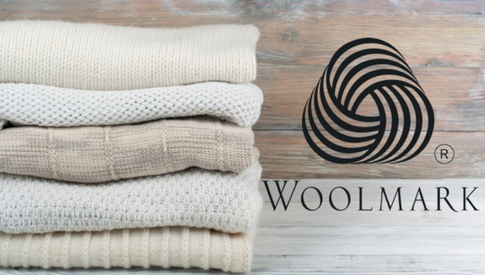 Stos złożonych swetrów w odcieniach beżu i bieli obok logo Woolmark na drewnianym tle. Logo przedstawia czarny, spiralny wzór z napisem \'Woolmark\'.