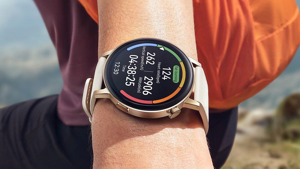 Na zdjęciu widoczny jest smartwatch z okrągłym wyświetlaczem, na którym pokazane są dane dotyczące aktywności fizycznej, takie jak liczba kroków i spalonych kalorii. Urządzenie jest noszone na nadgarstku osoby ubranej w pomarańczową koszulkę.