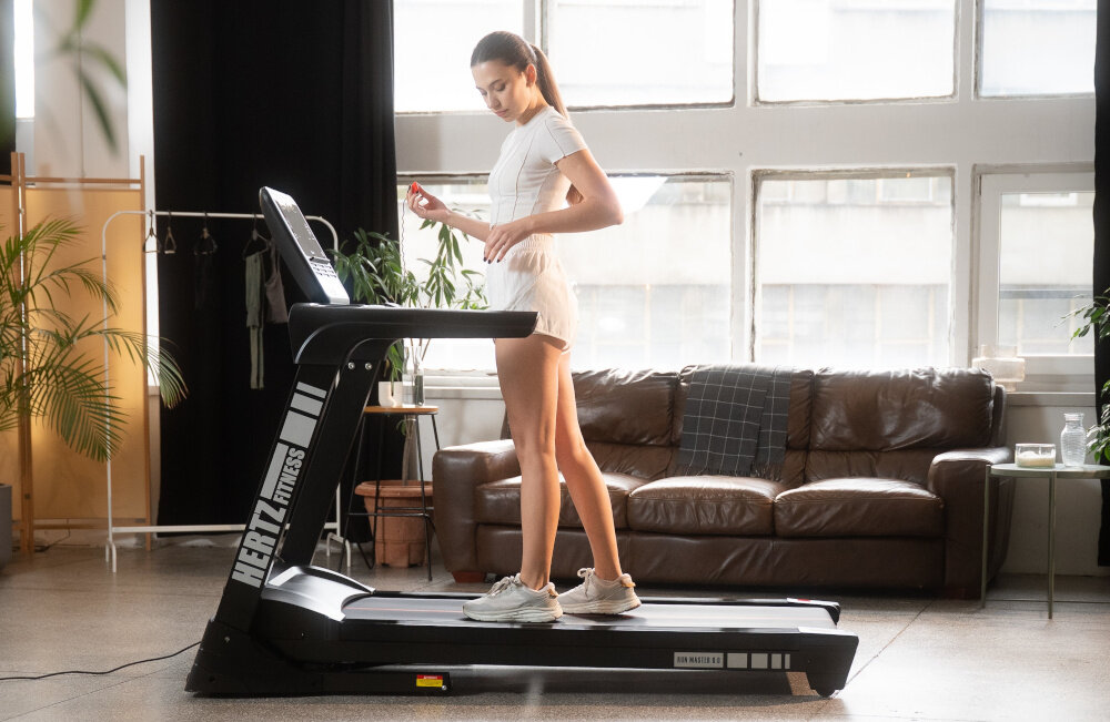 Osoba ćwiczy na bieżni elektrycznej z widocznym logo \'HERTZ FITNESS\'. W tle znajduje się sofa i duże okna, a obok bieżni stoją rośliny doniczkowe.