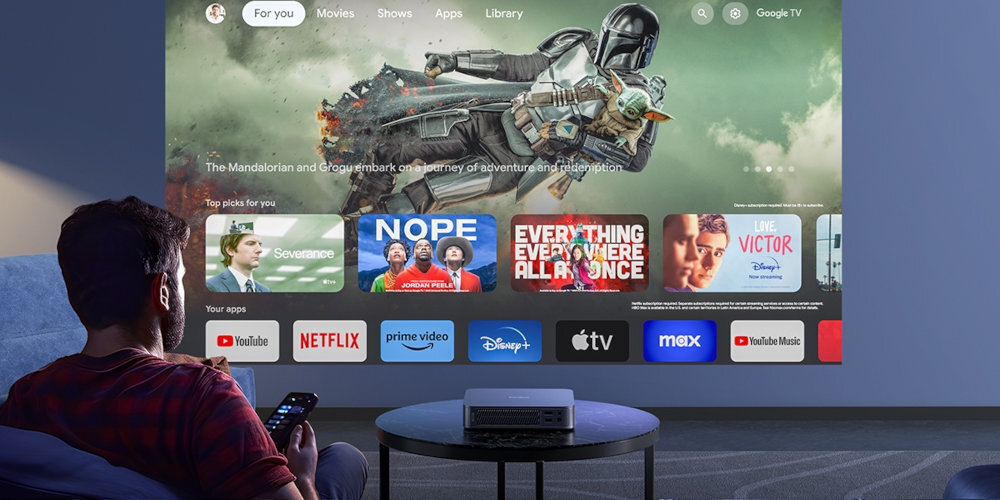 Osoba siedzi na kanapie, trzymając pilot, przed nią znajduje się projektor wyświetlający interfejs z aplikacjami takimi jak YouTube, Netflix, Prime Video, Disney+, Apple TV, Max i YouTube Music. Na ekranie widoczny jest obraz z filmu \'The Mandalorian and Grogu embark on a journey of adventure and redemption\'.