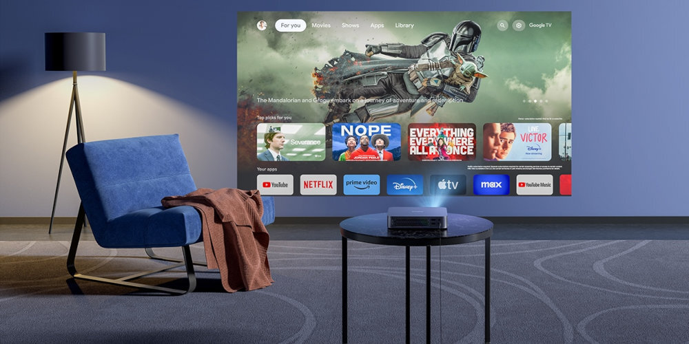 W pokoju na stoliku znajduje się projektor wyświetlający obraz na ścianie z interfejsem aplikacji streamingowych, takich jak Netflix, YouTube, Prime Video i Disney+. Obok stoi niebieski fotel z narzuconym kocem, a w tle widoczna jest lampa stojąca.