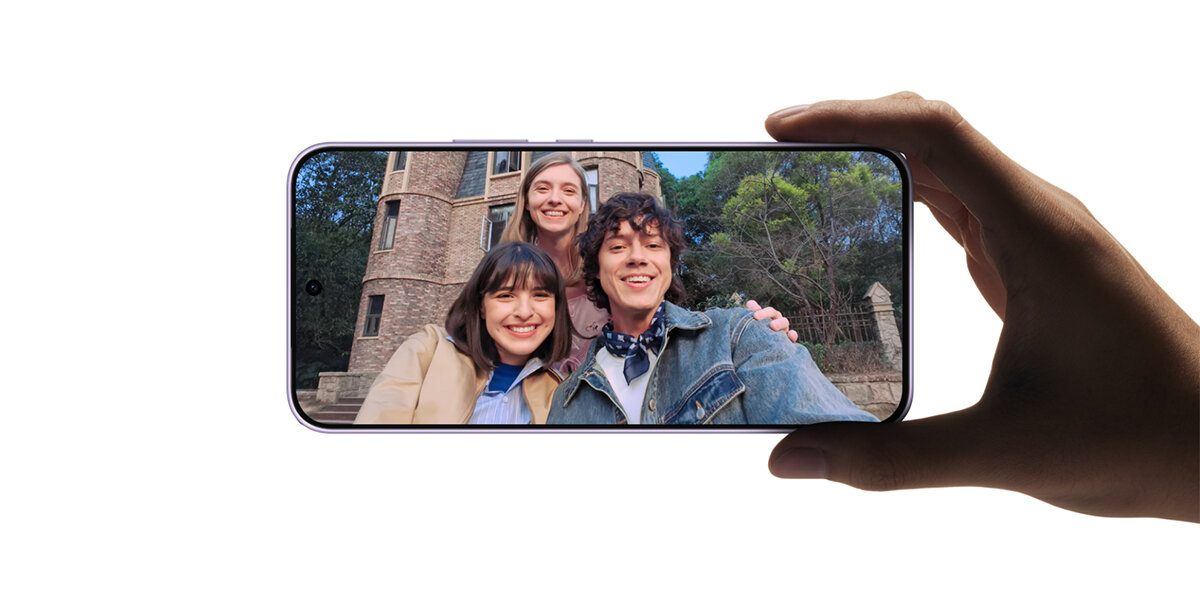 Osoba trzyma smartfon, na ekranie którego widoczne jest zdjęcie trzech uśmiechniętych osób na tle budynku i drzew.