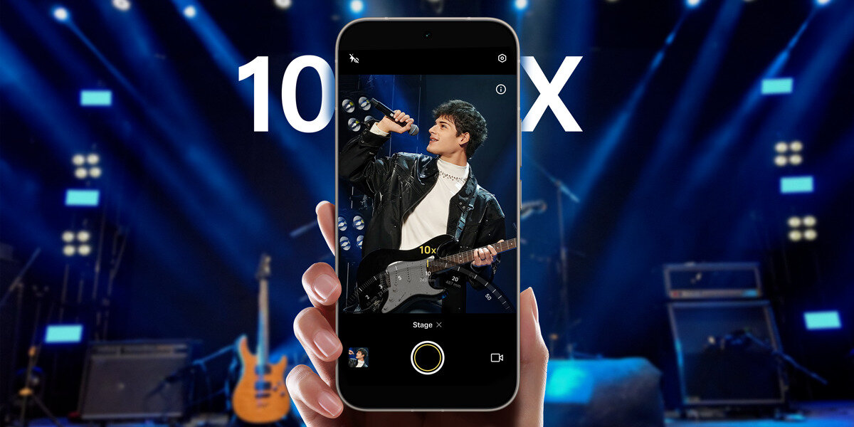 Osoba trzyma smartfon, na ekranie którego widać zdjęcie mężczyzny z gitarą na scenie. W tle sceny widoczne są światła i sprzęt muzyczny, a na ekranie telefonu napis \'10x\'.