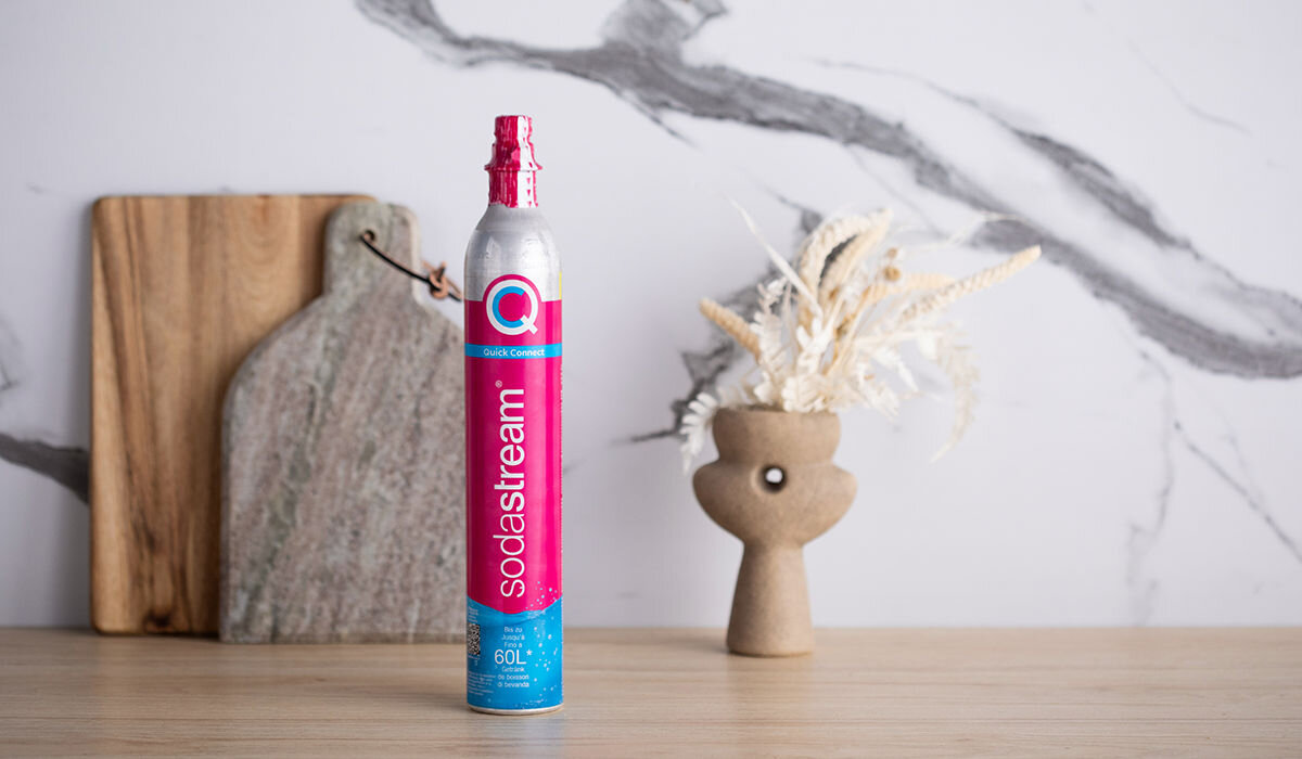 Na drewnianym blacie stoi metalowy cylinder z różową etykietą z napisem \'sodastream\'. W tle widoczne są deski do krojenia i wazon z suszonymi roślinami.