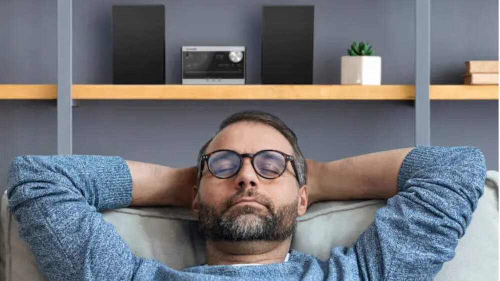 Mężczyzna w okularach siedzi na kanapie z zamkniętymi oczami, z rękami za głową. Na półce za nim znajduje się wieża audio z dwoma głośnikami i małą rośliną w doniczce.